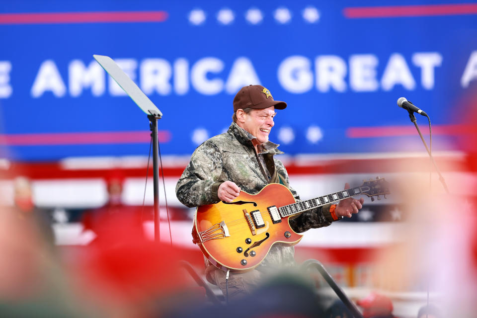 Musician in camouflage jacket playing an electric guitar at a rally with "Make America Great Again" sign in background