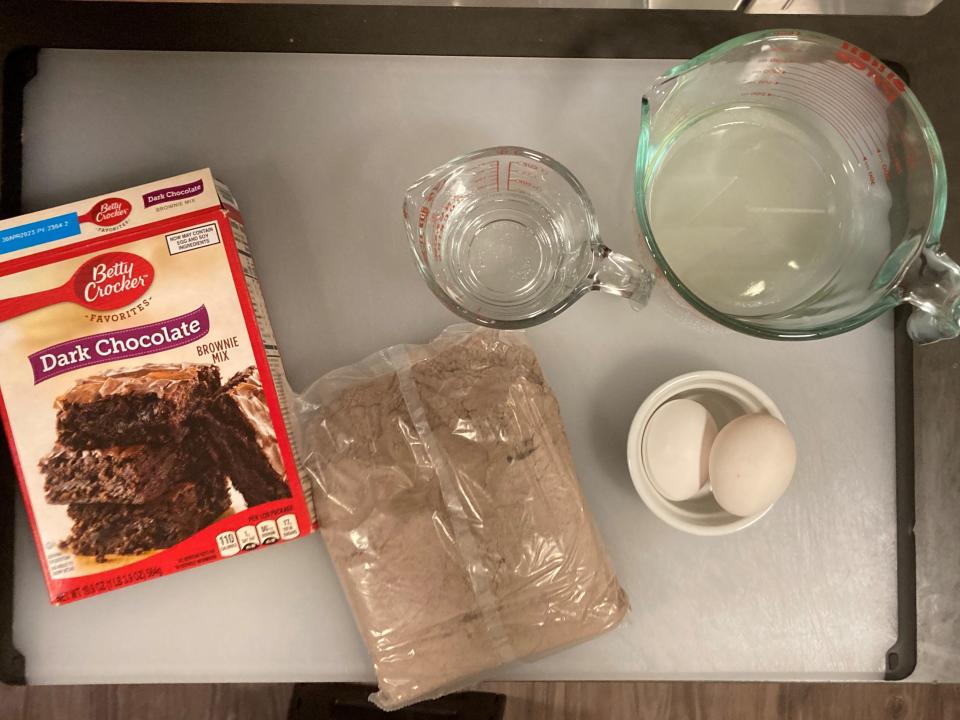 Betty Crocker Dark Chocolate brownie mix, brownie mix in package, bowl with two eggs, and liquid measuring cups with oil and water on a cutting board