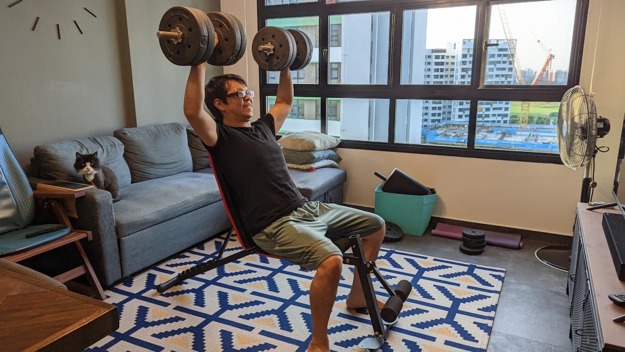 A Chinese man lifting weights on a sit-down bench in his living room home gym while his cat hangs around. (Photo: Yahoo Lifestyle Singapore)