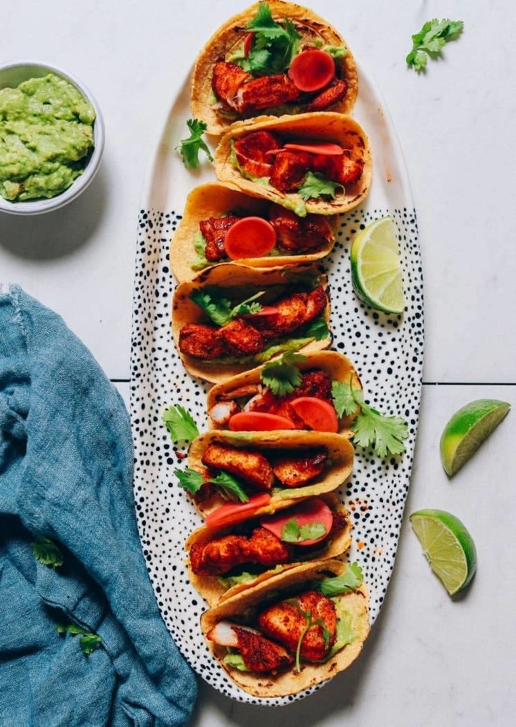 Plate of fish tacos garnished with cilantro, a side of guacamole in a bowl, and lime wedges on a table