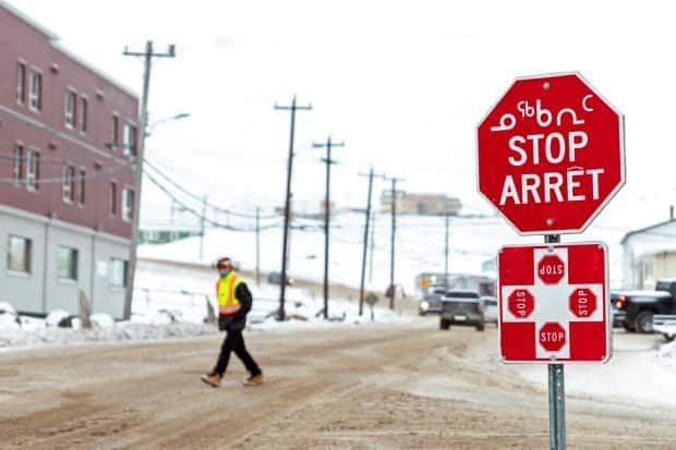 A man wearing a mask to help slow the spread of coronavirus disease (COVID-19) passes a stop sign written in English, French and Inuktitut. There were 74 active cases of COVID-19 in Nunavut as of Saturday. (Natalie Maerzluft/Reuters - image credit)