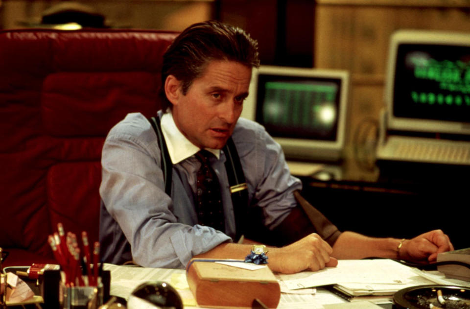 Michael Douglas, in a business suit with rolled-up sleeves, is seated at a cluttered desk with computers and papers in a scene from "Wall Street"