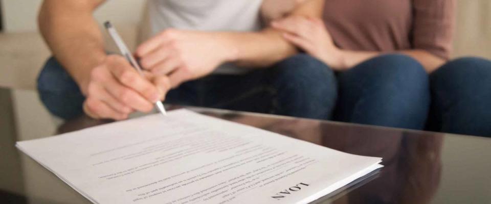 Close up of signing loan agreement, couple sitting on sofa, male hand with a pen setting signature, taking bank credit with easy terms of payment and low interest rate for purchasing property