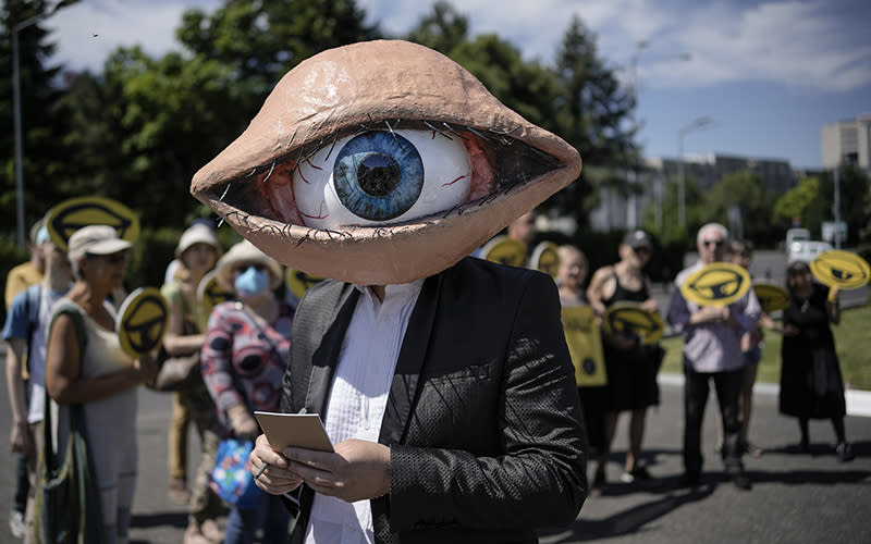 An activist wears a mask depicting a large eyeball as people hold yellow eyeball signs in the background