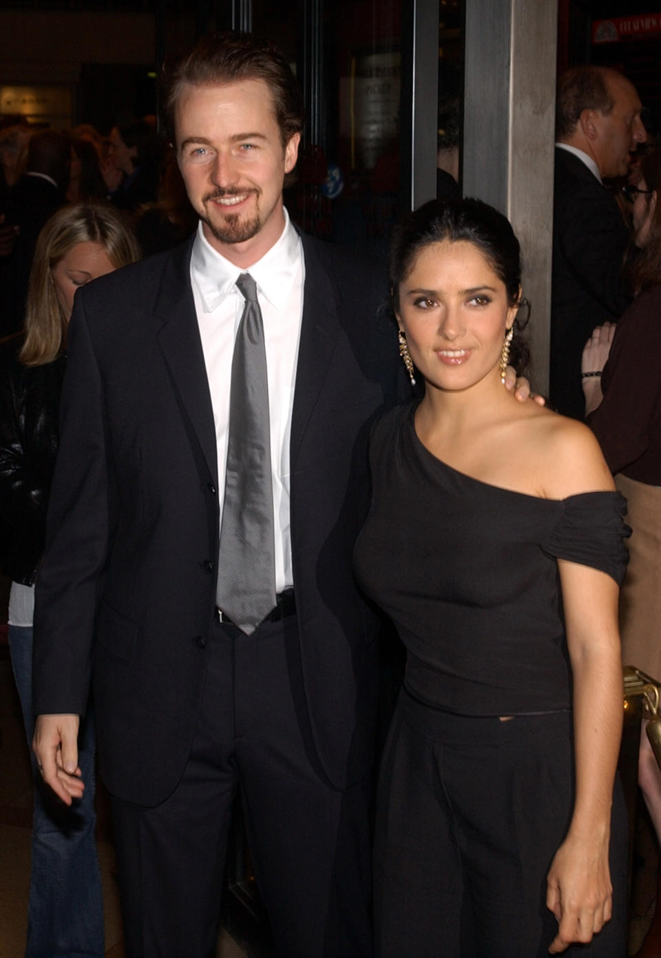 Edward Norton, wearing a suit and tie, stands with his arm across Salma Hayek's shoulder. Salma is wearing a two-piece outfit and her hair pulled back