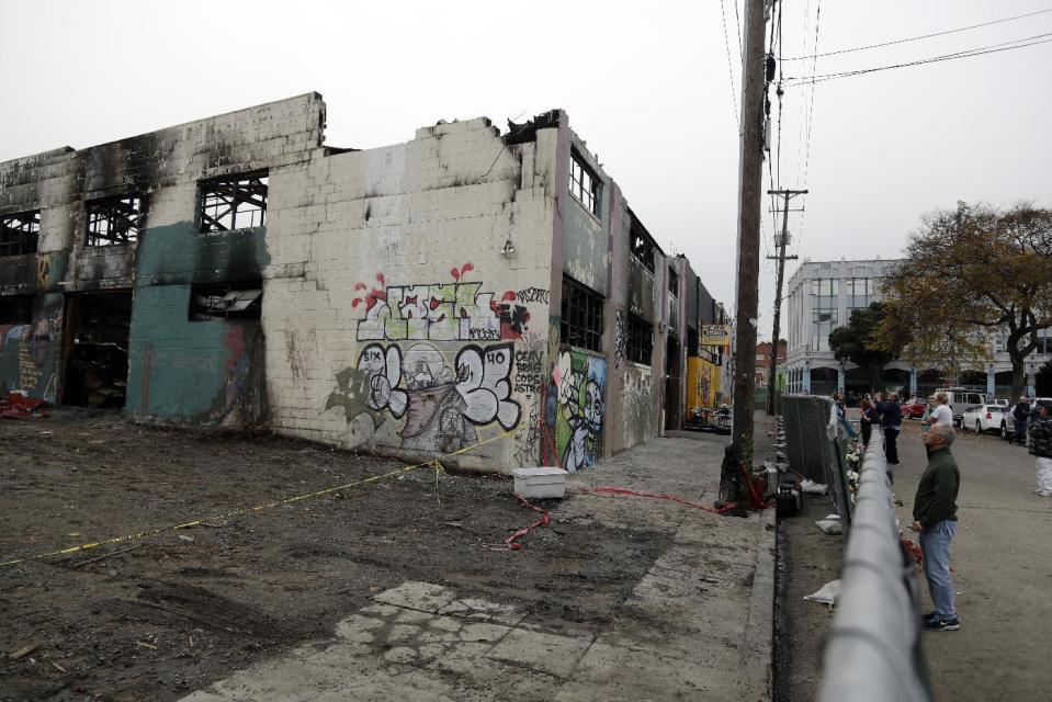 People look at the scene of a warehouse fire from behind a fence Tuesday, Dec. 13, 2016, in Oakland , Calif. The fire killed dozens of people during a electronic dance party as it raced through the building, in the deadliest structure fire in the U.S. in more than a decade. (AP Photo/Marcio Jose Sanchez)