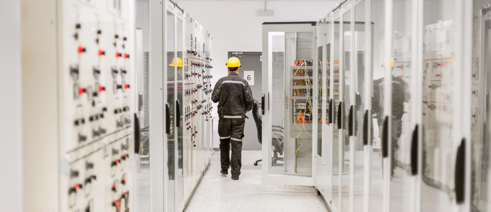 An employee in safety clothing and yellow hard hat walks through a bank of switches in a building's engineering room.