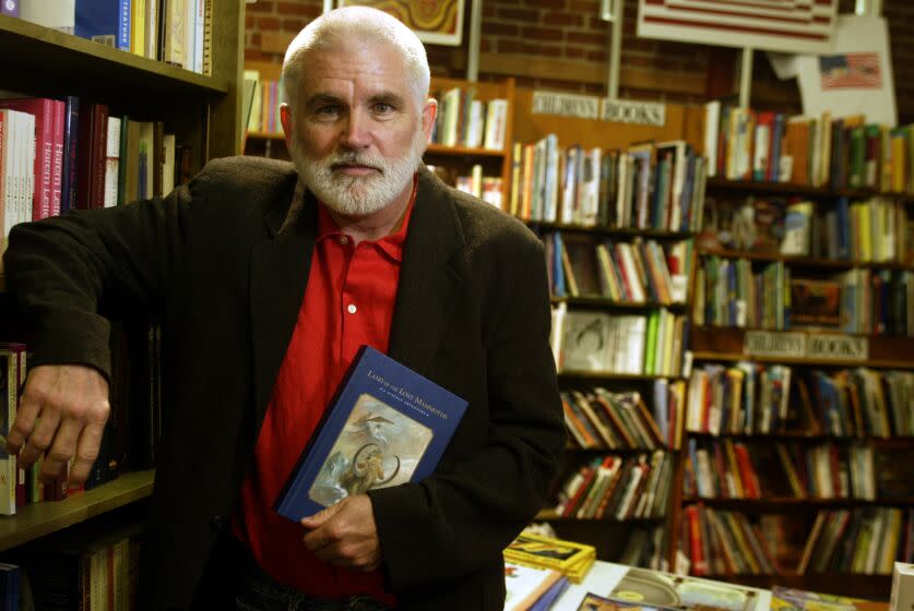 Mike Davis, in a red shirt and brown blazer, holds a book while leaning on a shelf inside a book shop