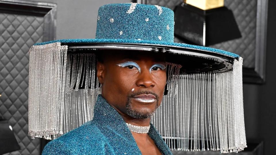 A Black man wears makeup and a blue hat with lampshade-esque fringe