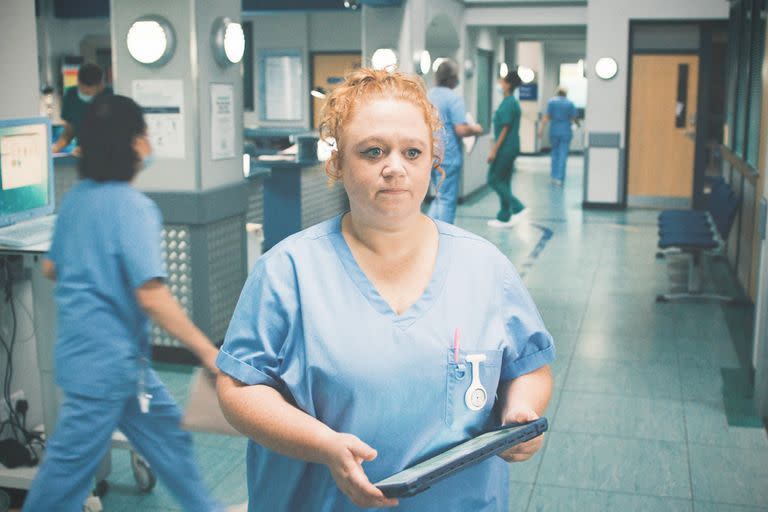 robyn miller in casualty, a woman stands in the middle of a hospital corridor with people walking around her