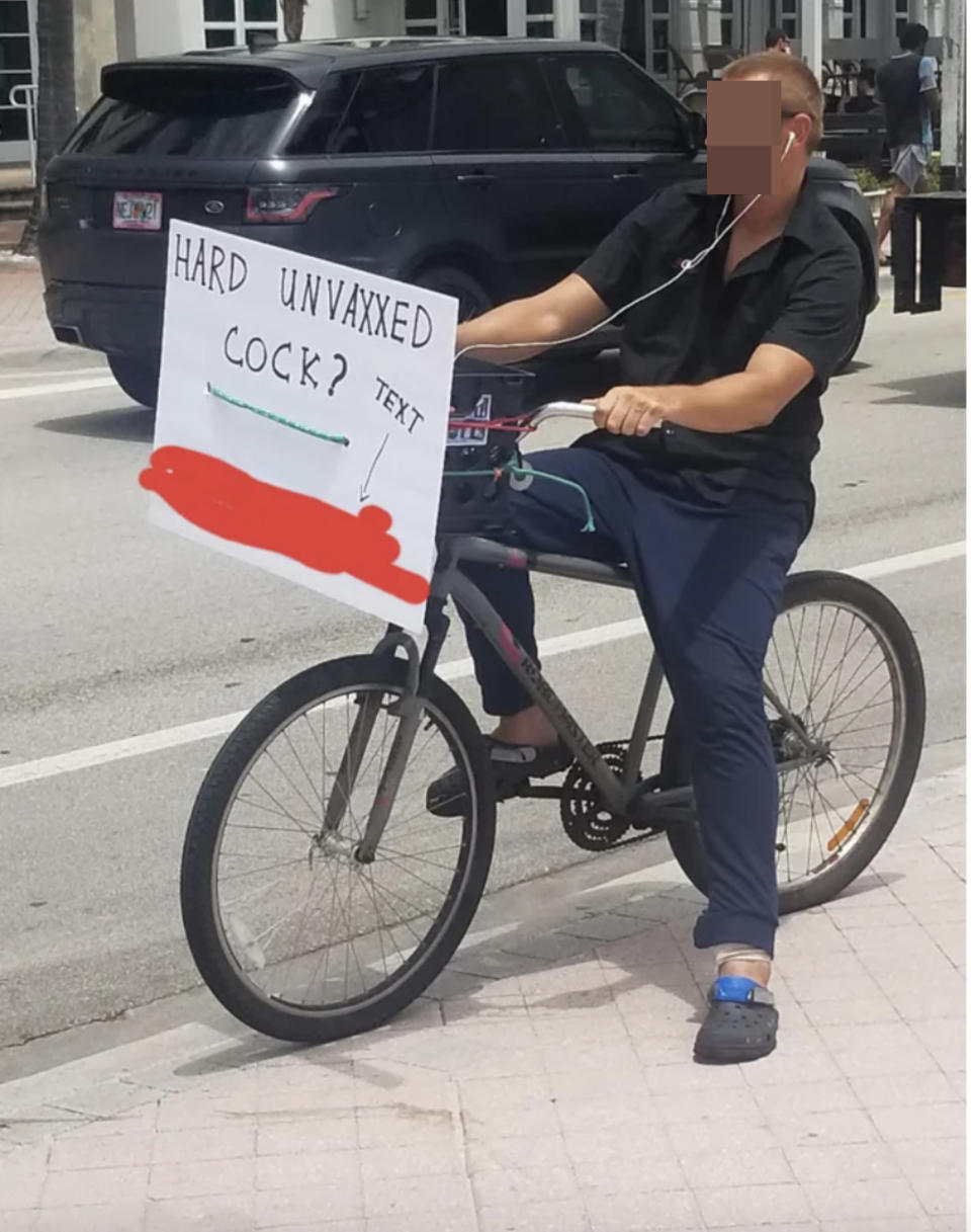 a man riding a bike with a sign that says, "Hard unvaxxed cock — text," and a phone number