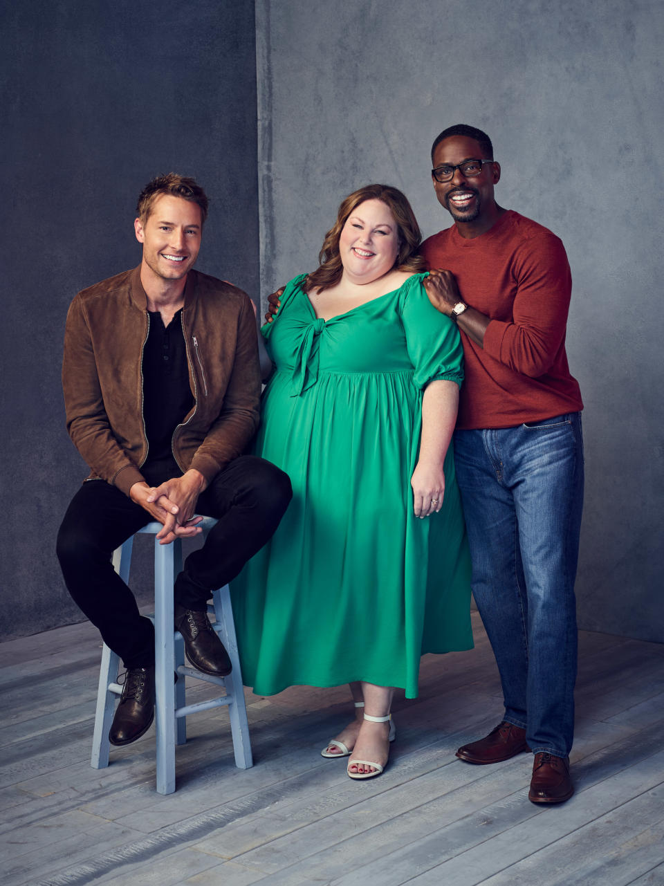 Justin Hartley, Chrissy Metz, and Sterling K. Brown pose together. Justin sits on a stool, wearing a casual jacket; Chrissy wears a flowy dress; Sterling wears a sweater and jeans