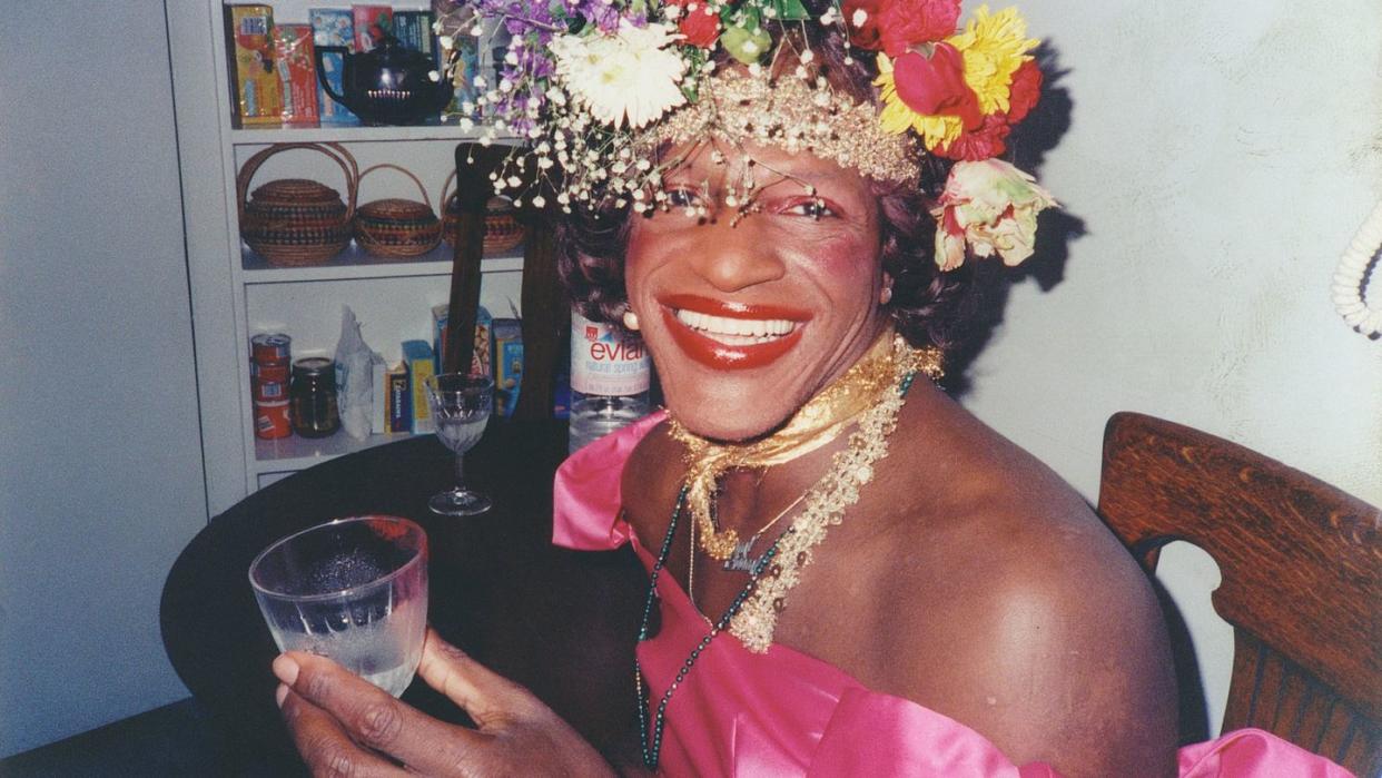 marsha p johnson smiles at the camera while seated at a table, she wears a flower crown and a bright pink dress and holds a glass