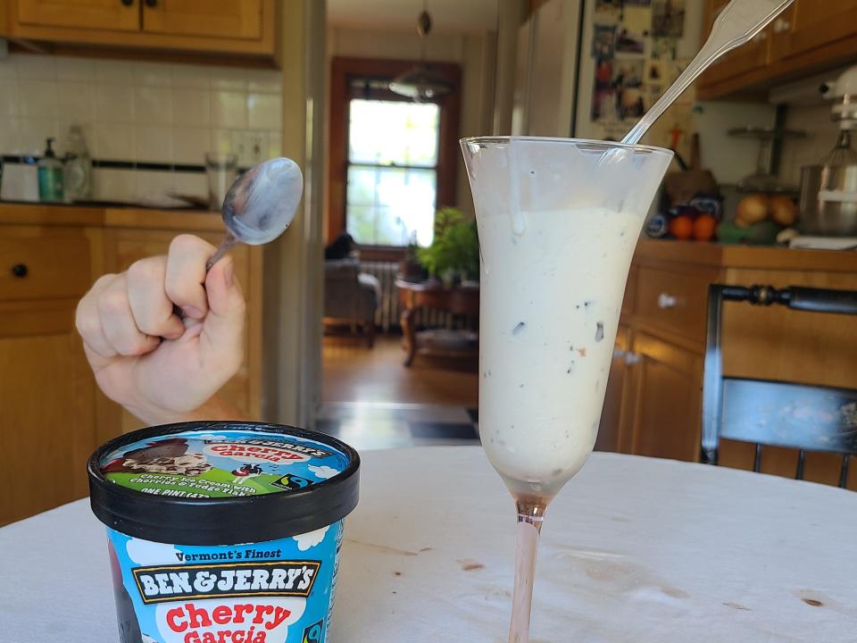 A hand holding up a spoon behind the table with the carton and serving of ice cream