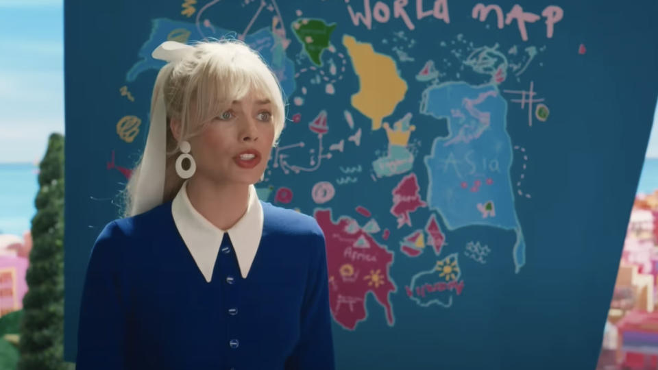 “Barbie,” dressed in a blue dress with a white collar, stands in front of a map of the world showing the disconnected shapes of different geographical areas.