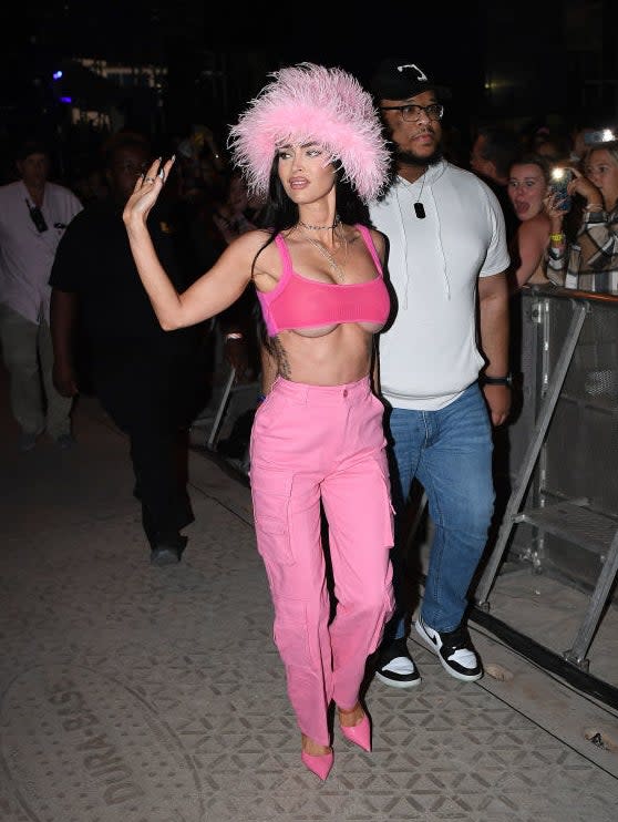 Cardi B in a pink outfit with a feathered hat, waving, with a bodyguard and fans in the background