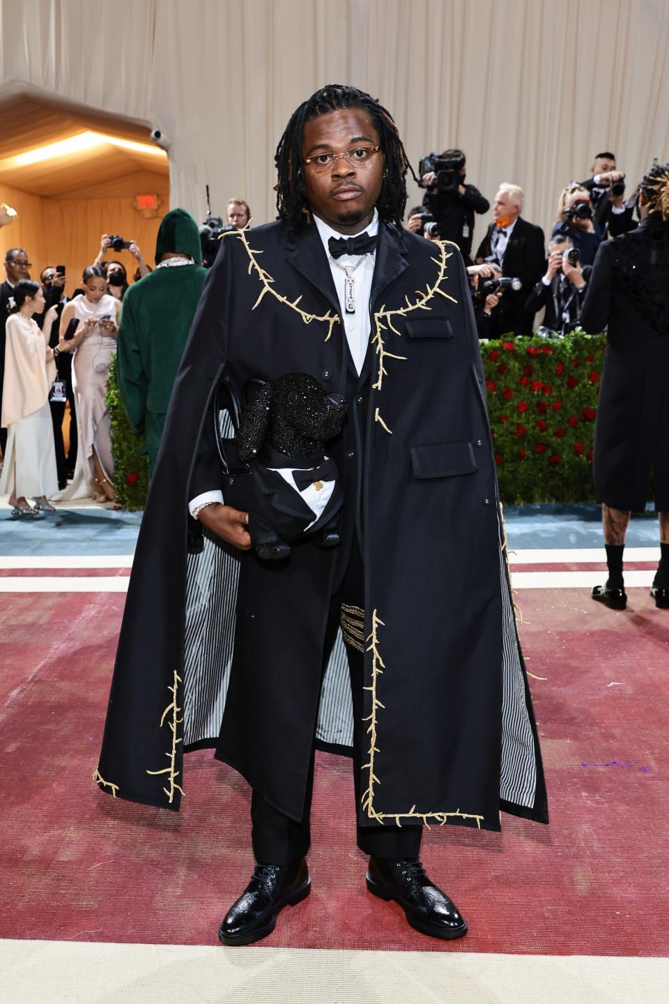 Gunna in a black suit and black cape decorated with strands of gold thorn branches. He's holding a crystal black dog purse in a tuxedo.