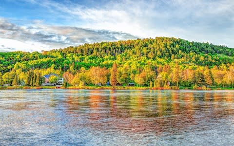 Chicoutimi river in Saguenay, Quebec, with riverfront houses and forest during sunset with colorful foliage reflection - Credit: iStock