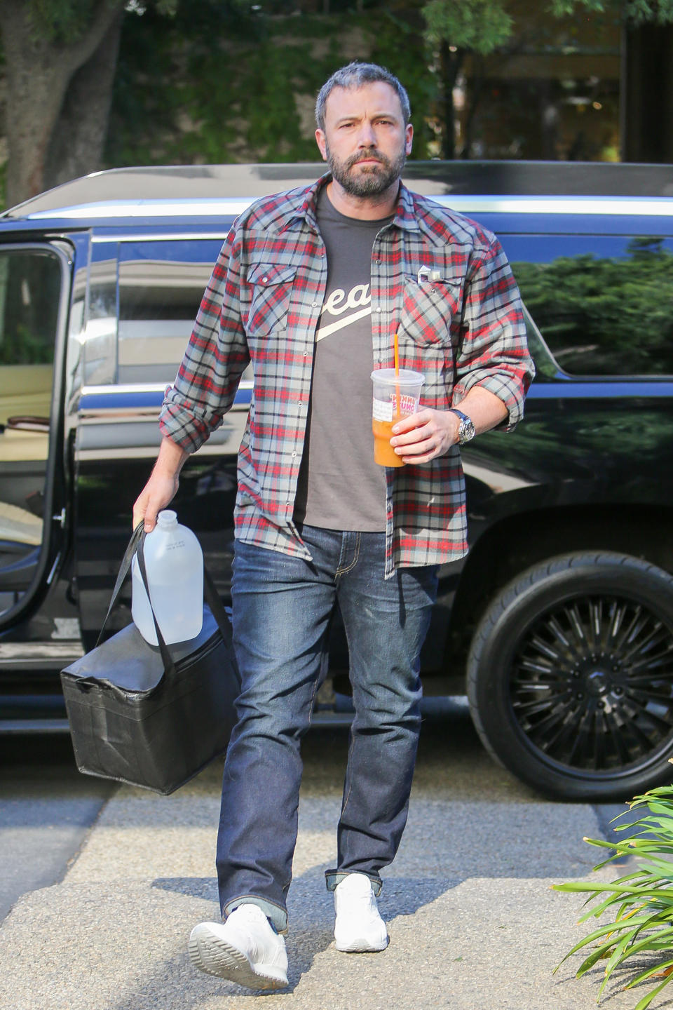 Man in a plaid shirt over a graphic tee, denim, and sneakers, carrying a beverage and a bag, exiting a vehicle