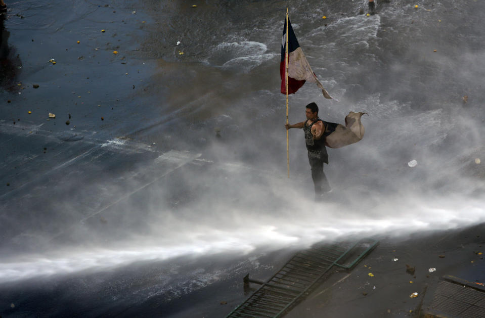 Holding a Chilean flag, a protester braces against a water canon sprayed from a police truck amid a march by students and union members in Santiago, Chile, Monday, Oct. 21, 2019. Protesters defied an emergency decree and confronted police in Chile’s capital on Monday, continuing disturbances that have left at least 11 dead and led the president to say the country is “at war.” (AP Photo/Miguel Arenas)