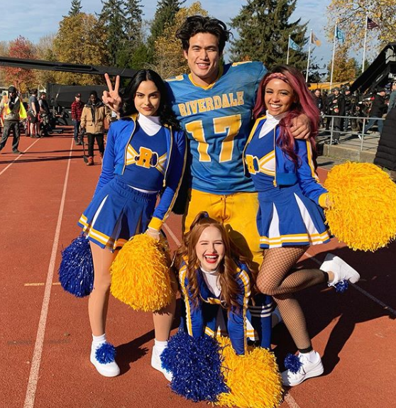 A photo of Camila Mendes, Charles Melton, Madelaine Petsch and Vanessa Morgan wearing yellow and blue sports uniforms on the set of Riverdale.