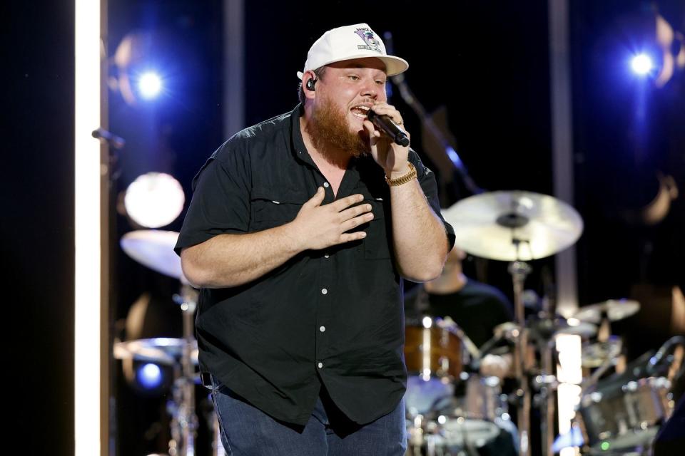 luke combs on a stage singing into a microphone, standing in front of a set of drums, wearing a black shit, jeans, and a white baseball cap