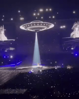 Concert stage with a lit central structure, crowd in darkness, and large screens displaying a performer