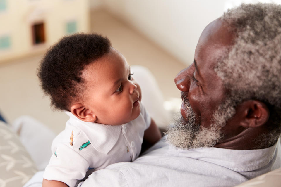 An elderly man with a beard holds and gazes at a baby who is looking back at him. Both appear happy and engaged