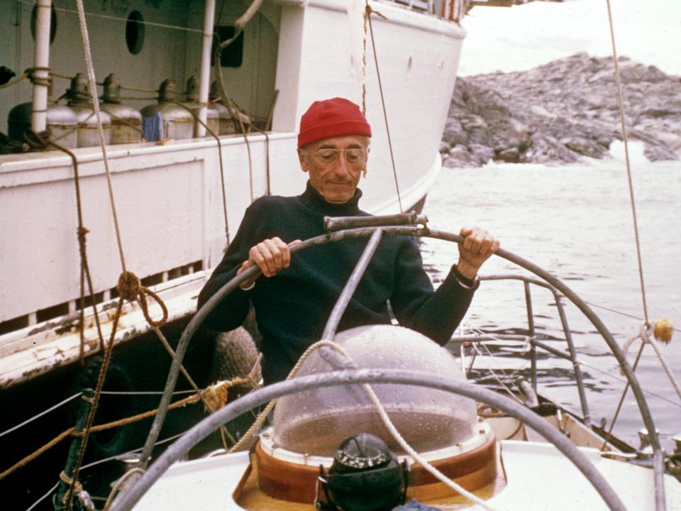 Jacques Cousteau wears a red hat and steers his boat, Calypso.