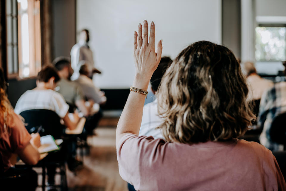 Person raising hand in a seminar with attendees and speaker in background