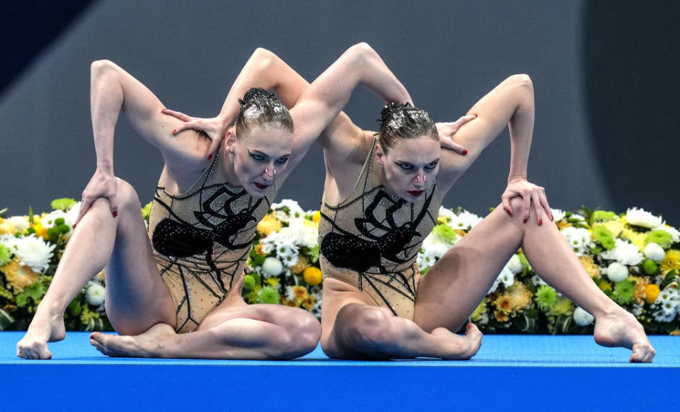 Two swimmers with arms and legs bent, one arm resting on their own knee and the other resting on the other's shoulder