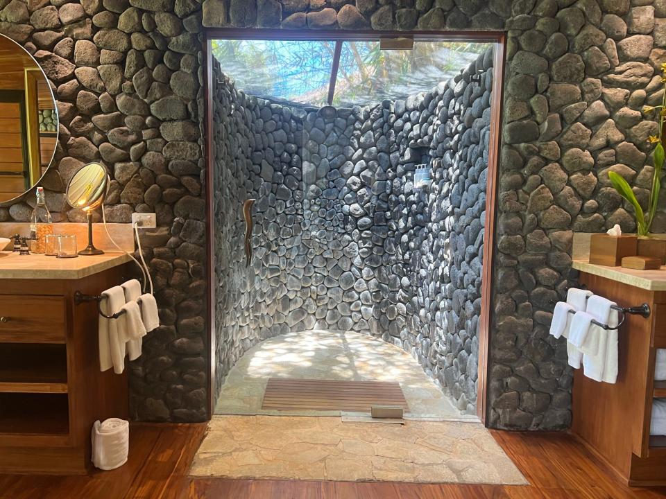 Lava-rock walkout shower with trees and sky over the top of the shower. The shower is connected to a bathroom with wooden details