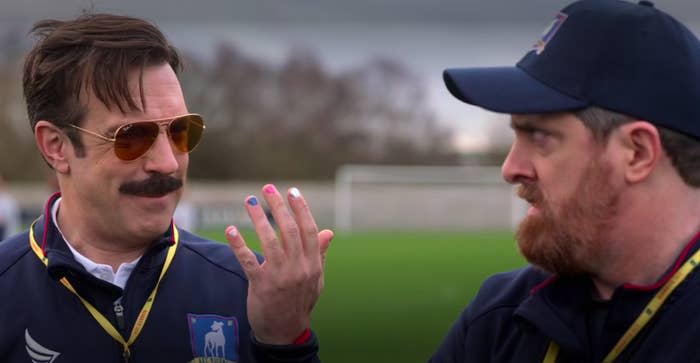 Ted Lasso showing his hand with different-colored fingernails to coach Beard