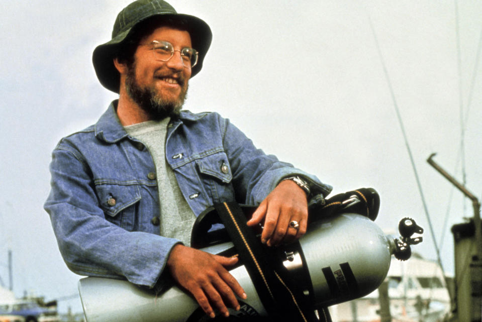 Richard Dreyfuss as Matt Hooper holding scuba gear in a scene from the film Jaws. He is smiling and wearing glasses, a hat, and a denim jacket