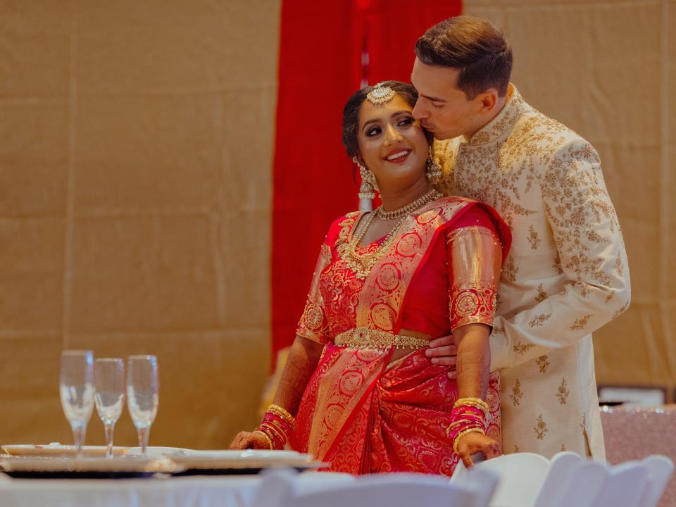 The couple are in a reception hall wearing traditional Indian wedding clothing. The groom is stood behind the bride, giving her a kiss on the cheek while she smiles at him.