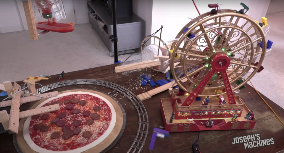 A wacky pizza-making robot uses its cheese-coneA pizza machine using its ferris wheel to pour olives on a mid-production pizza.