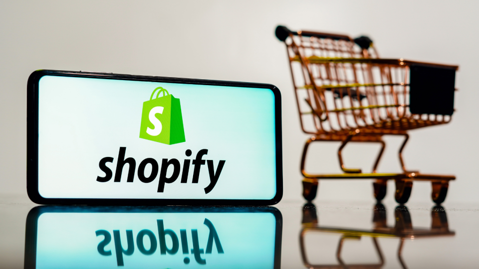 Shopify logo on phone with shopping cart in the background.