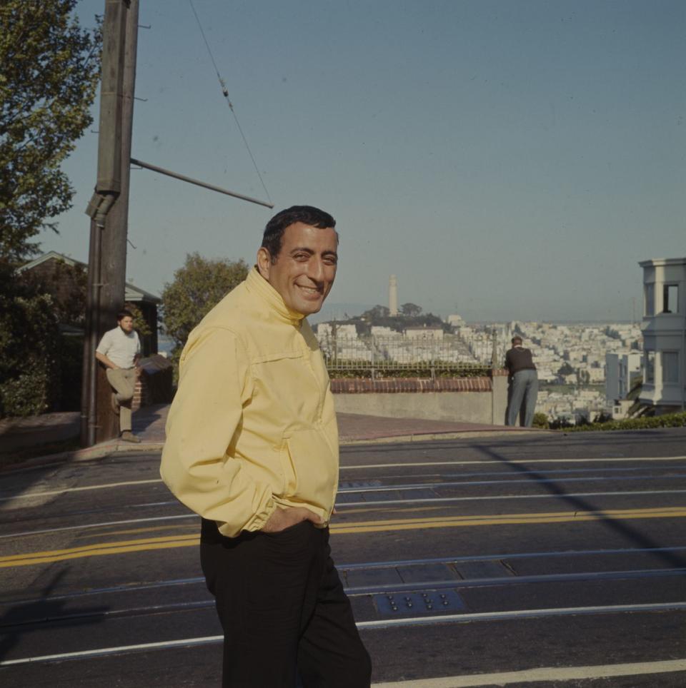 tony bennett smiles at the camera while walking on a street with his hands in his pants pockets, he wears a yellow jacket and black pants, behind him is a view of coit tower and surrounding buildings in san francisco