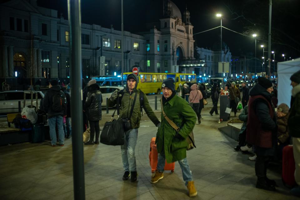 Two men in hats and coats, carrying or rolling bags, are seen on a city street.