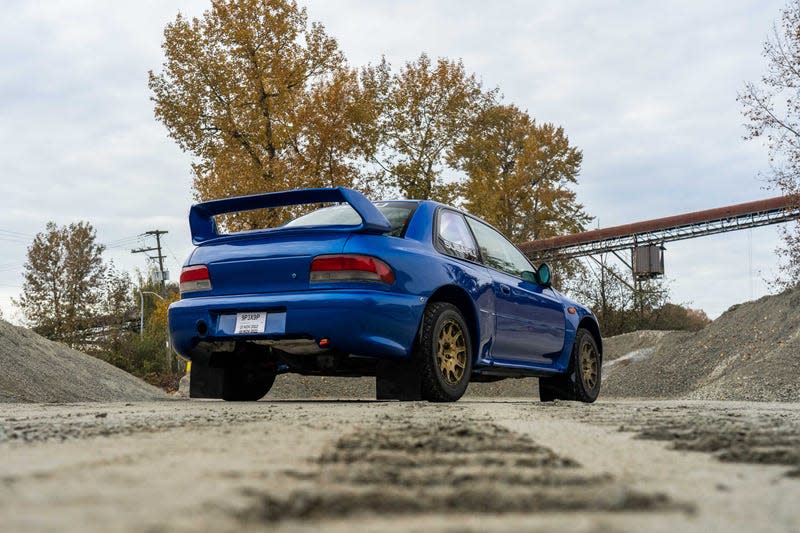 rear shot of a blue subaru coupe parked on a gravel lot. the photo is taken from an extremely low vantage point, showing off the rally-racing rear spoiler on the subaru's trunk lid, as well as the gold wheels and all-terrain tires