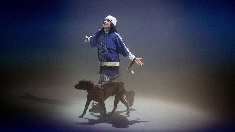 Billie Eilish on stage for a listening party for her album Hit Me Hard and Soft in LA in May. Billie is a 22-year-old woman with long dark hair. She wears a white cap twisted backwards and a blue sports shirt. She's on stage with her dog, Shark. 