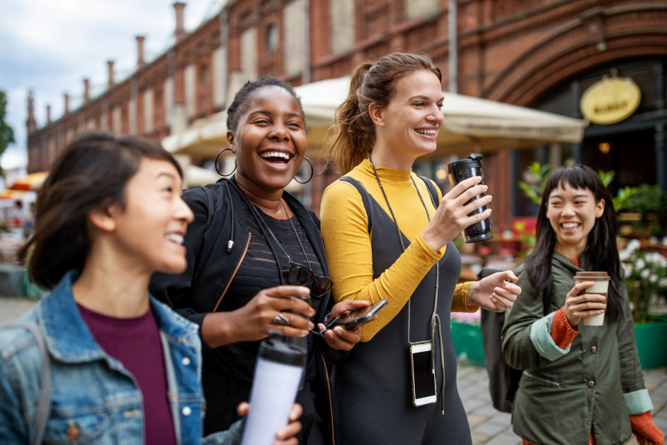 Four friends laugh and walk outdoors with coffee cups