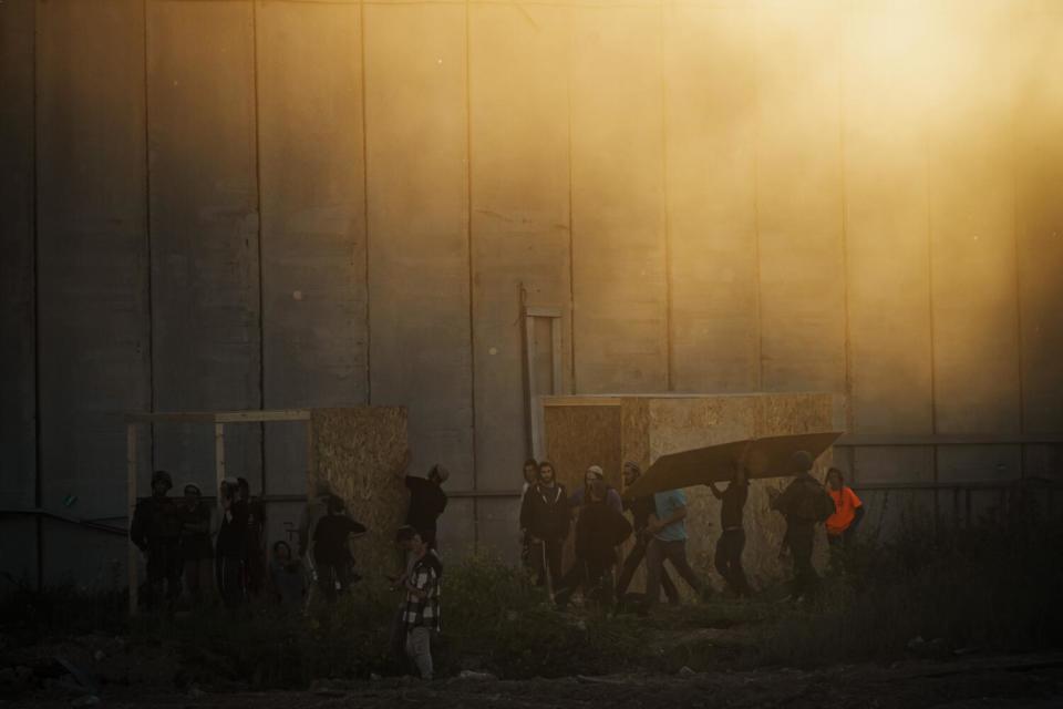 Over a dozen people building small structures in front of a tall wall in the haze