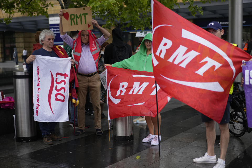 Workers and members of the RMT (The National Union of Rail, Maritime and Transport Workers) union hold up banners and placards as they pose for photographs whilst standing in the rain on a picket line outside King's Cross station, in London, during a railway workers strike, Thursday, June 23, 2022. Tens of thousands of railway workers walked off the job in Britain on Tuesday, bringing the train network to a crawl in the country's biggest transit strike for three decades. (AP Photo/Matt Dunham)