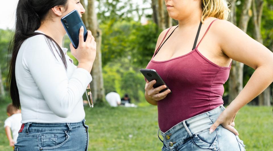 Two women standing in a park having a conversation. One is holding a phone to her ear, and the other is looking at her phone