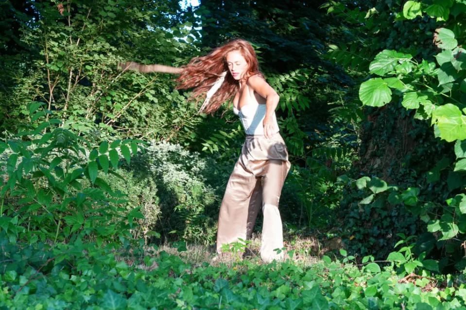 A woman with long red hair twisting her body, standing in nature