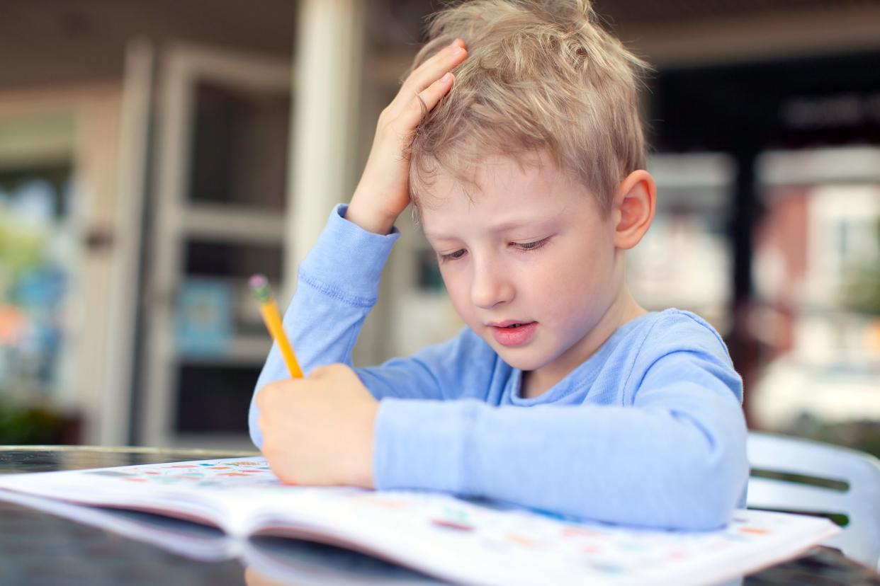 Focus on young boy concentrating while holding his head with his hand, holding a pencil on book while sitting at a student desk at elementary school with a blurred background of windows and door of classroom