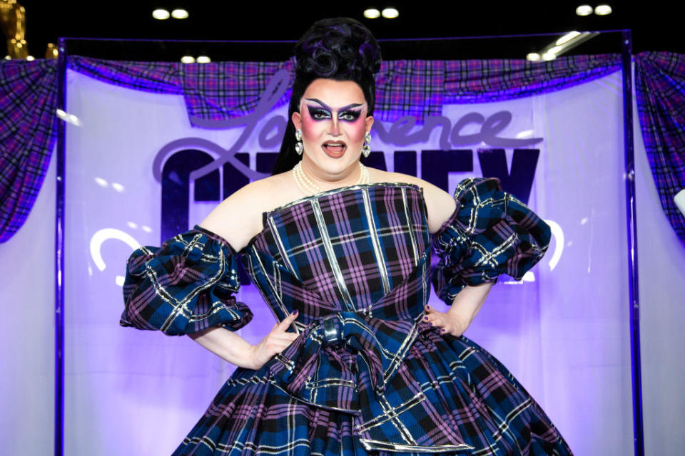 Drag queen Laurence Chaney in a voluminous plaid dress striking a confident pose at an event