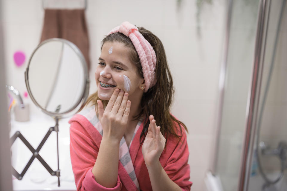 Retinol serum. Cheerful young girl wearing a bathrobe and makeup hairband when applying face cream in the bathroom