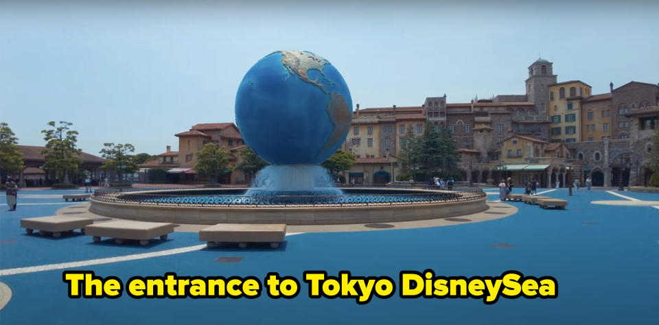 Fountain with a large globe centerpiece in a public square, surrounded by benches and buildings. There are a few people walking around